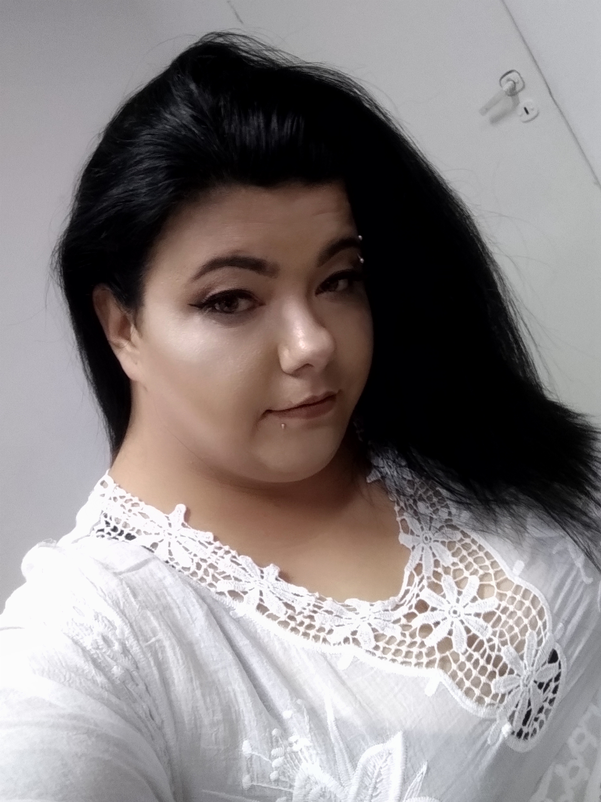 Portret kobiety z ciemnymi włosami, z delikatnym makijażem oczu i piercingiem na brodzie oraz nad brwią, ubrana w białą, koronkową bluzkę.