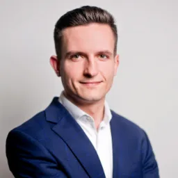 Portret młodego mężczyzny w niebieskiej marynarce w kratę i białej koszuli, uśmiechającego się do kamery na jasnym tle.