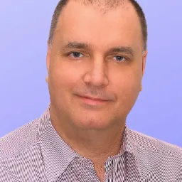 Portret mężczyzny w koszuli w paski na jasnoniebieskim tle. Mężczyzna ma krótkie włosy, niebieskie oczy i lekki uśmiech. Ujęcie od przodu, skupione na twarzy.