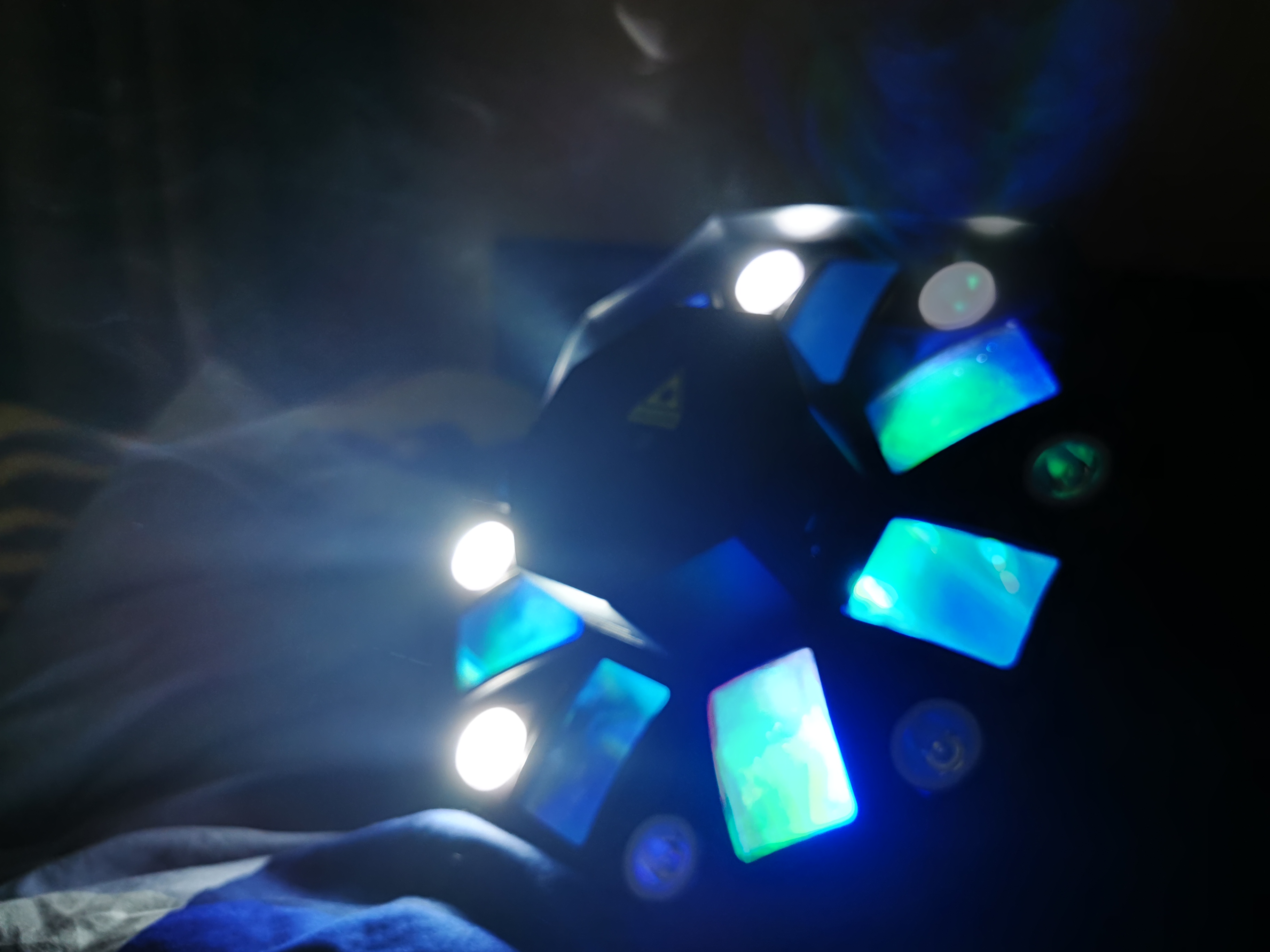 Wielobarwny reflektor dyskotekowy z białymi diodami i niebiesko-zielonymi panelami, leżący na pogniecionej tkaninie w ciemnym otoczeniu.