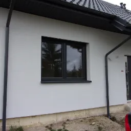 Fragment elewacji domu z oknem i rynnami w kolorze antracytowym, widoczne ocieplenie fundamentów i surowy teren wokół budynku.