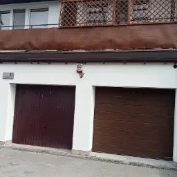 Dwukondygnacyjny budynek z dwoma brązowymi bramami garażowymi, białymi ścianami i balkonem z brązową osłoną oraz drewnianą kratą. Na ścianie widoczny numer domu i system alarmowy.