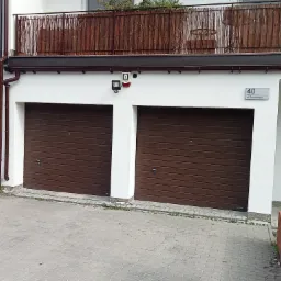 Biała elewacja budynku z dwoma brązowymi bramami garażowymi, adres '40 ul. Siewierzan' widoczny na ścianie, balkon z brązową matą, oświetlenie zewnętrzne i system alarmowy.