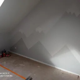 Ściana pod skosem w pokoju na poddaszu z motywem gór malowanym w dwóch odcieniach szarości, widoczne puszki z farbą i narzędzia malarskie.