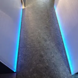 Nowoczesna podłoga z szarych płytek i listwami LED wzdłuż ścian, tworzącymi niebieskie oświetlenie akcentujące. Minimalistyczny design.