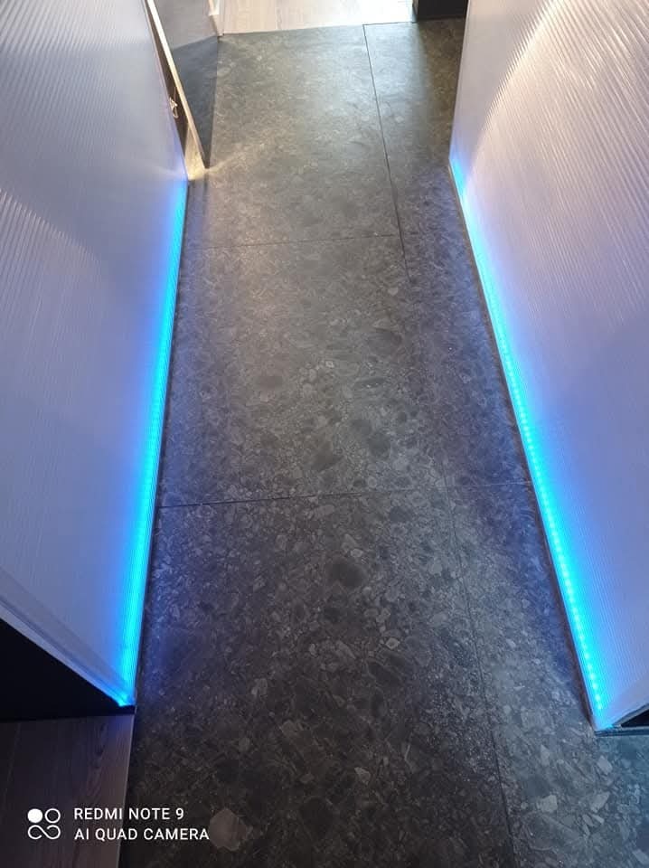 Nowoczesna podłoga z szarych płytek i listwami LED wzdłuż ścian, tworzącymi niebieskie oświetlenie akcentujące. Minimalistyczny design.