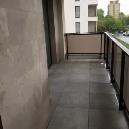 Wykończony balkon z szarymi płytkami na podłodze i ścianie, widok na budynek mieszkalny w tle, szklana balustrada z czarną ramą.