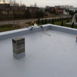 Płaski dach pokryty szarą membraną, z kominem z betonowych bloczków, narzędziami i materiałami budowlanymi w tle oraz widokiem na zabudowania mieszkalne.