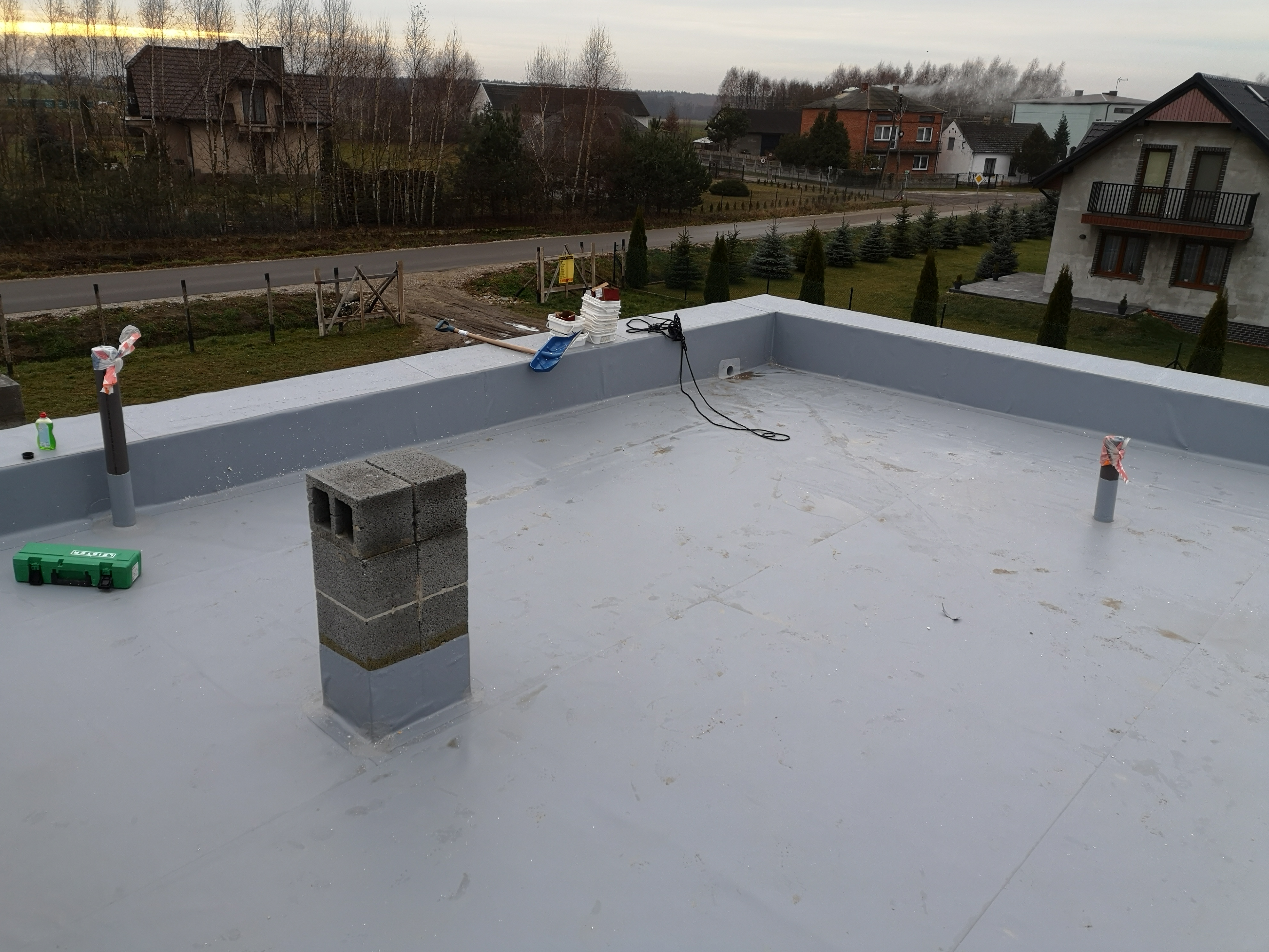 Płaski dach pokryty szarą membraną, z kominem z betonowych bloczków, narzędziami i materiałami budowlanymi w tle oraz widokiem na zabudowania mieszkalne.
