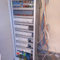 Otwarta szafa sterownicza z licznymi przewodami i zabezpieczeniami, widoczne kable w kolorach pomarańczowym, niebieskim i białym zwisające wzdłuż ściany, instalacja w trakcie prac.