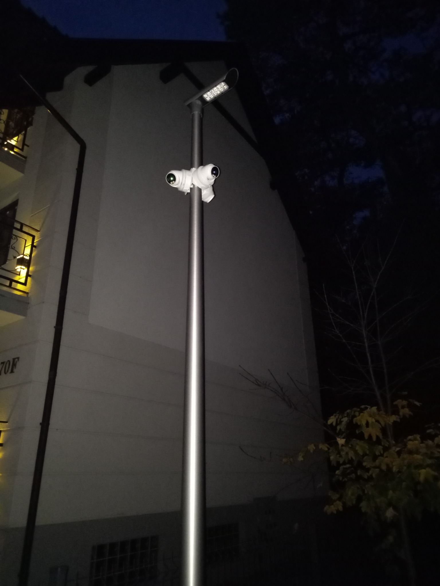 Dwie kamery przemysłowe zamontowane na metalowym słupie oświetleniowym przed budynkiem mieszkalnym, nocne ujęcie.