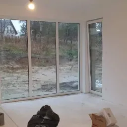 Wnętrze świeżo wykończonego pokoju z dużymi oknami tarasowymi, widoczne ślady po pracach wykończeniowych, drabina malarska i kartony na podłodze.