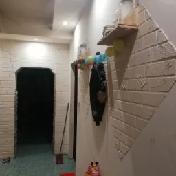 Korytarz z dekoracyjną ścianą z imitacji białej cegły, drewnianymi półkami z słoikami i kolorowymi lampionami, widoczne zabawki dziecięce, mop i niedokończone przejście do innego pokoju.