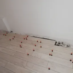 Układanie podłogi z paneli imitujących drewno, z użyciem systemu poziomowania płytek z czerwonymi klinami, widoczne niedociągnięcia przy ścianie.