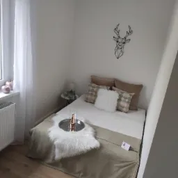 Przytulna sypialnia w odcieniach bieli i beżu z dekoracyjnym geometrycznym reniferem na ścianie, srebrną tacą z butelką i kieliszkami na ł&oacute;żku oraz futrzanym dywanikiem.