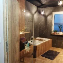 Nowoczesna łazienka z wanną obudowaną drewnem, szklaną kabiną prysznicową, ciemnymi płytkami imitującymi marmur i drewnianą podłogą ułożoną w jodełkę, oświetlona lampami wiszącymi o geometrycznych...