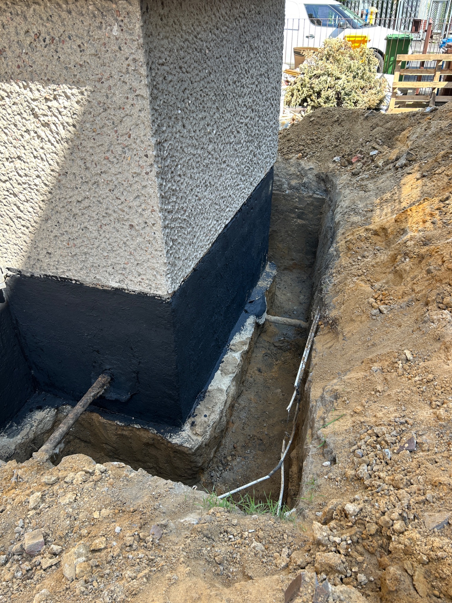 Fundament budynku zabezpieczony czarną izolacją, wykopany rów wokół. Widoczne przewody i fragmenty instalacji w wykopie. Tło: elementy otoczenia budowy.