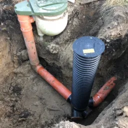 Wykonany drenaż rozsączający w wykopie, widoczne rury pomarańczowe łączące studzienkę z filtrem oraz rurę karbowaną pionową z pokrywą, ziemia wokół wykopu.