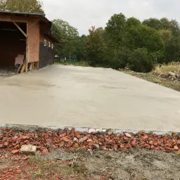 Wylany beton na placu budowy, widoczne pozostałości po cegłach i fragment budynku gospodarczego w tle.
