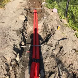 Wykop z ułożonymi dwoma czerwonymi rurami karbowanymi, widoczna ziemia, fragment ogrodzenia i koparka w tle.