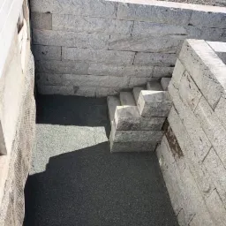 Kamienna konstrukcja schodów w zagłębieniu terenu, otoczona murem oporowym z szarego kamienia, wypełniona drobnym żwirem.