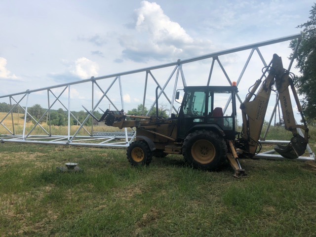 Żółto-czarna koparko-ładowarka marki Case z uniesionym ramieniem kopiącym, stoi obok długiej, metalowej konstrukcji kratownicowej na trawiastym terenie. W tle widoczne drzewa i pochmurne niebo.
