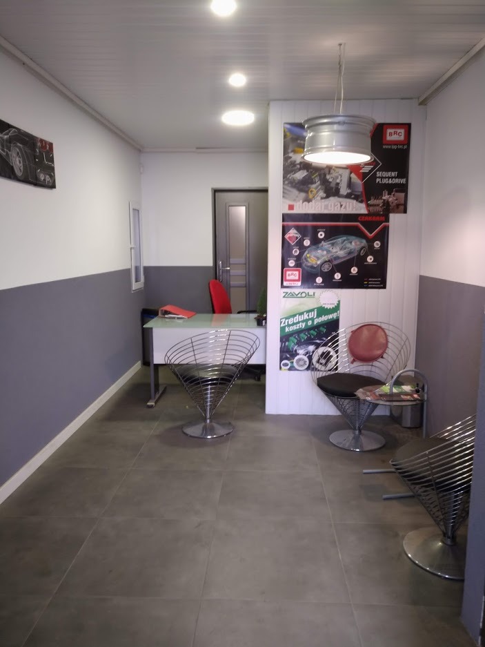 Poczekalnia w warsztacie samochodowym z designerskimi drucianymi krzesłami, biurkiem i plakatami BRC i Zavoli na ścianie, oświetlona lampą wykonaną z felgi samochodowej.