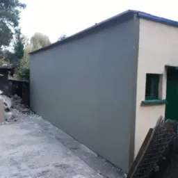 Świeżo otynkowana ściana budynku gospodarczego w kolorze szarym, widoczna zielona stolarka okienna i drzwiowa, na ziemi gruz i resztki materiał&oacute;w budowlanych.