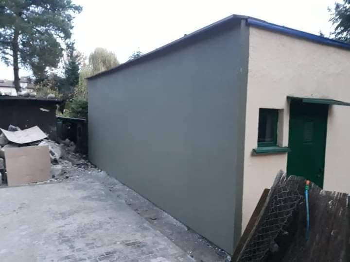 Świeżo otynkowana ściana budynku gospodarczego w kolorze szarym, widoczna zielona stolarka okienna i drzwiowa, na ziemi gruz i resztki materiałów budowlanych.