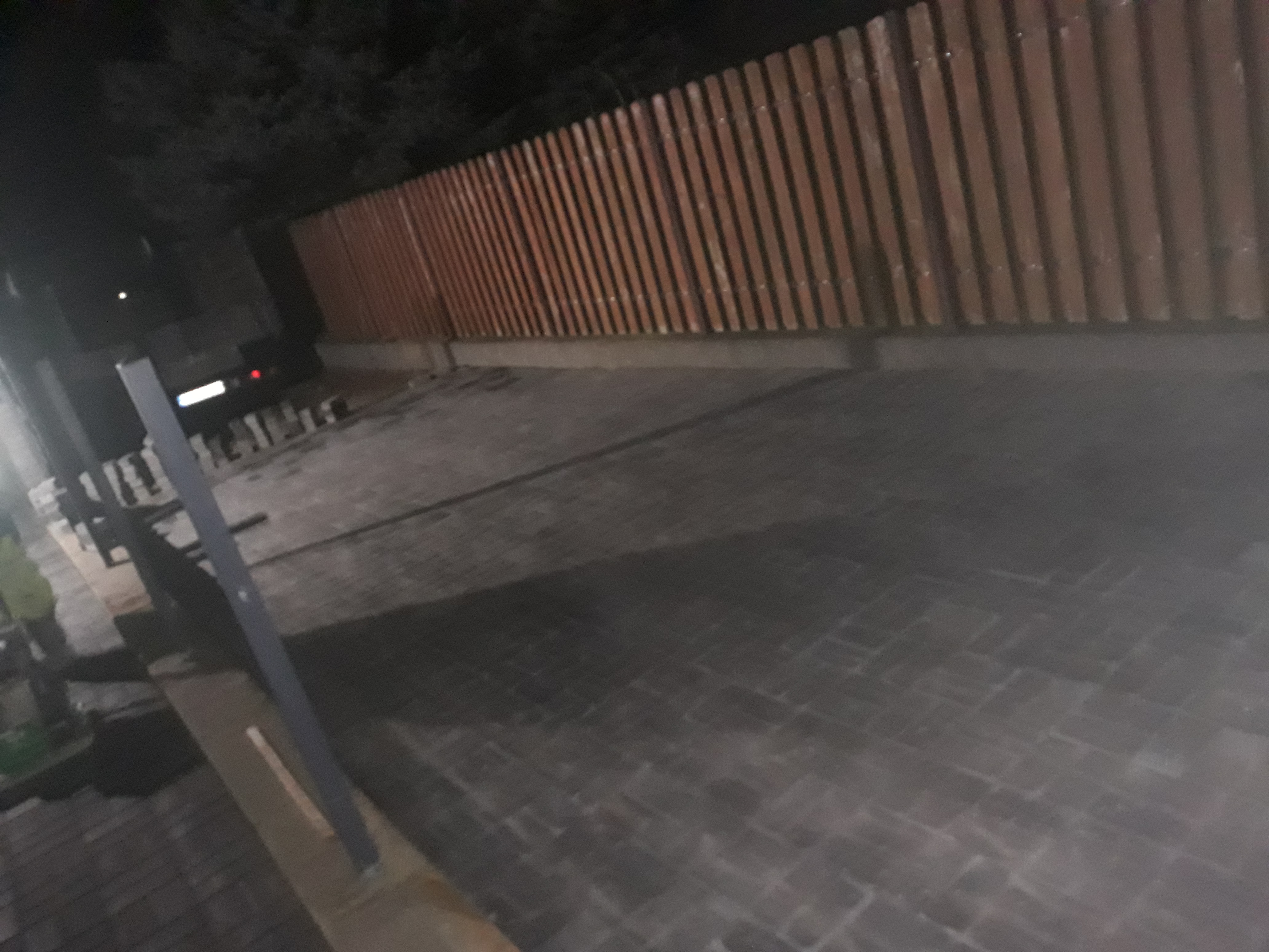 Ułożona kostka brukowa w odcieniach szarości, oświetlona nocą, z widocznym fragmentem drewnianego ogrodzenia w tle.