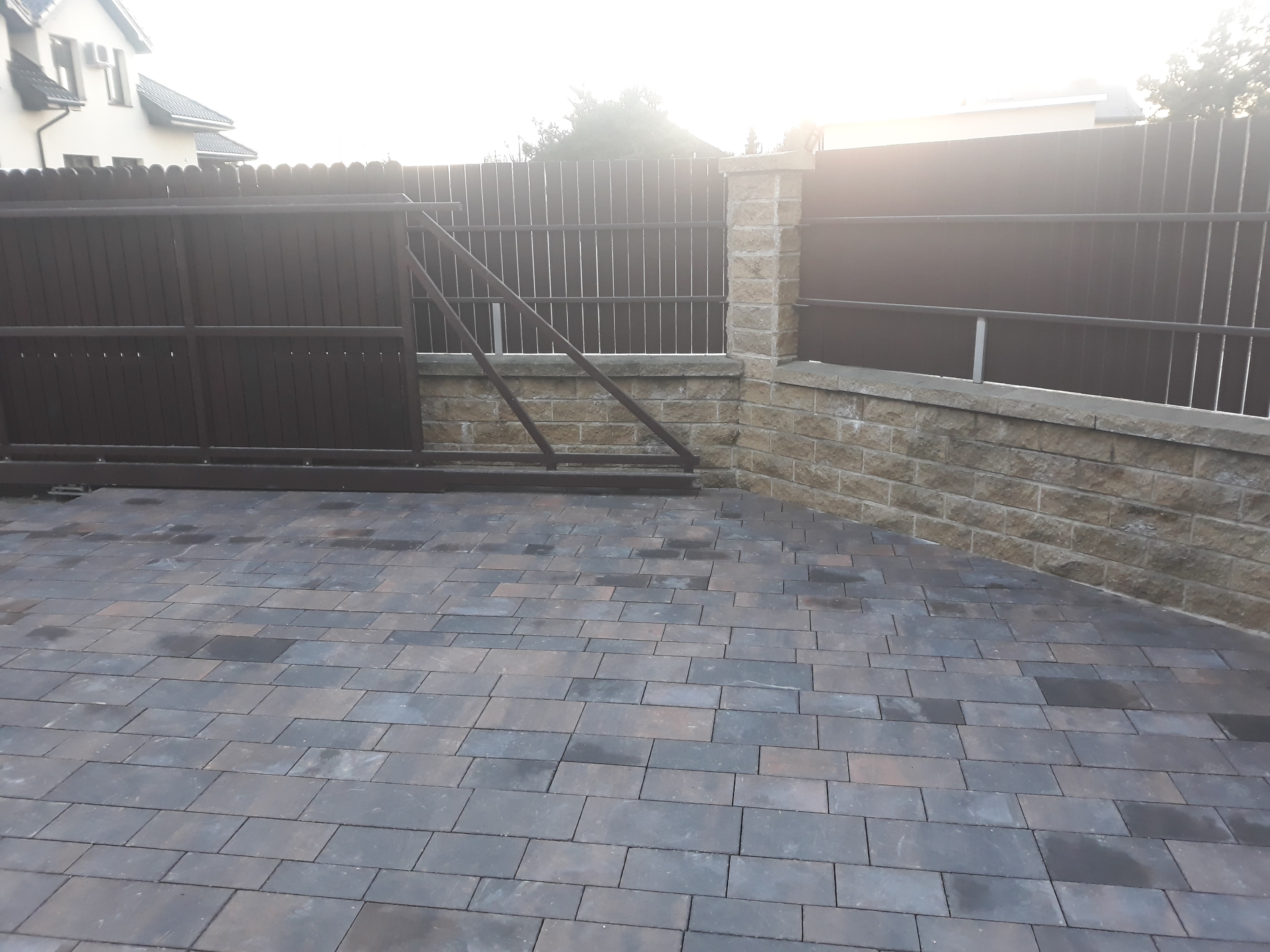 Ułożona kostka brukowa w odcieniach szarości i brązu, obok przesuwnej bramy z ciemnego drewna i fragmentu ogrodzenia z metalowymi prętami, wspartego na niskim murku z kamienia łupanego.