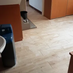 Ułożona podłoga z płytek imitujących drewno w trakcie remontu, widoczny filtr do wody BWT, wiadro i narzędzia glazurnicze w tle.