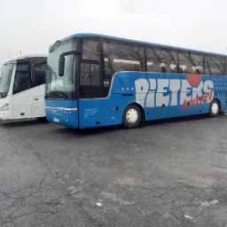 Niebieski autokar z białym napisem 'PIETEXS travel' zaparkowany obok białych autokarów na placu w pochmurny dzień.