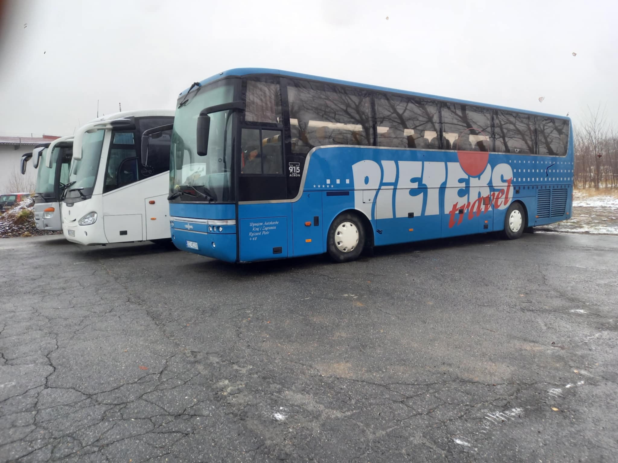 Niebieski autokar z białym napisem 'PIETEXS travel' zaparkowany obok białych autokarów na placu w pochmurny dzień.