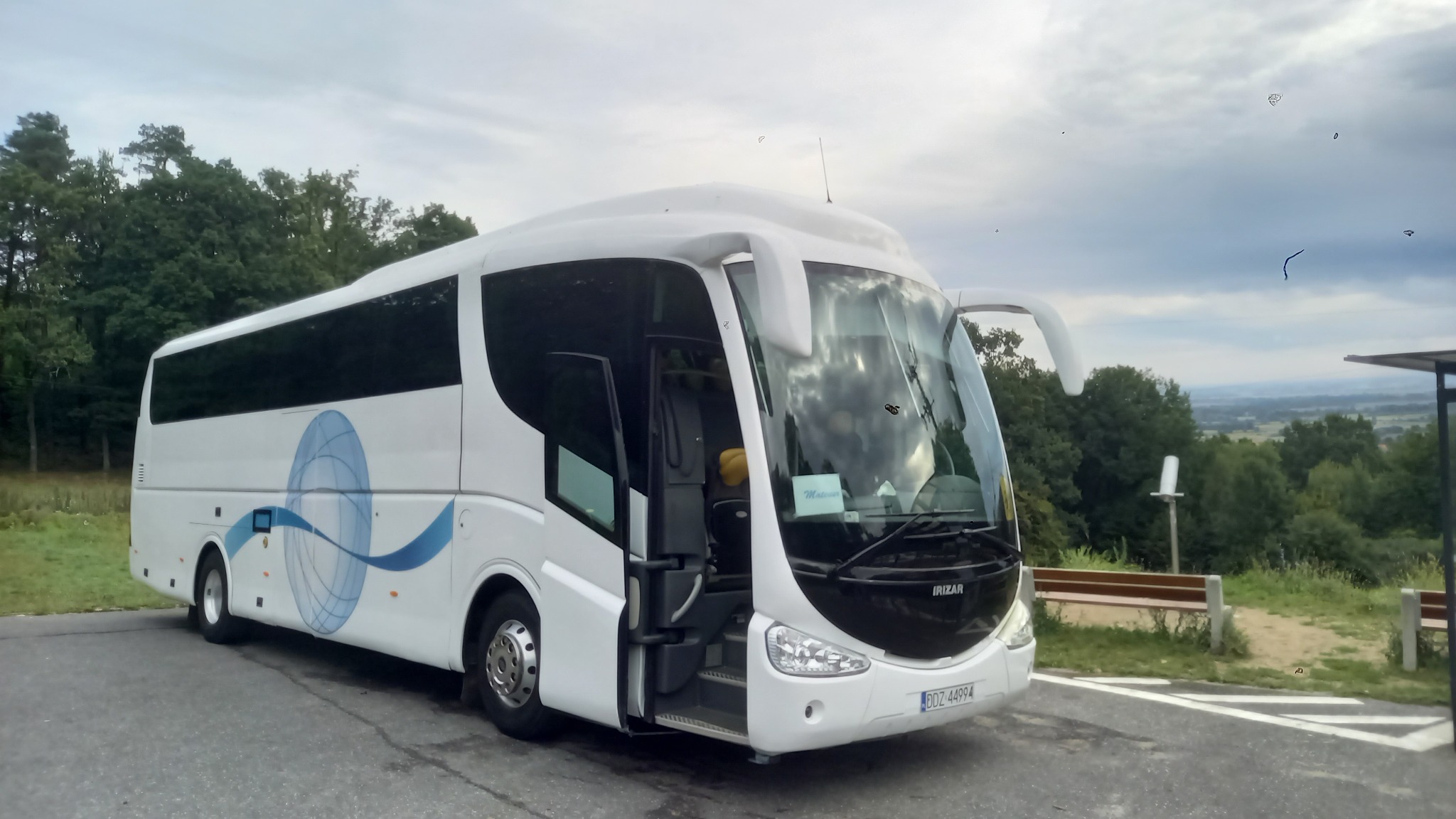 Biały autokar Irizar z otwartymi drzwiami na tle zieleni i pochmurnego nieba. Na szybie widoczna kartka. Autobus stoi na parkingu.