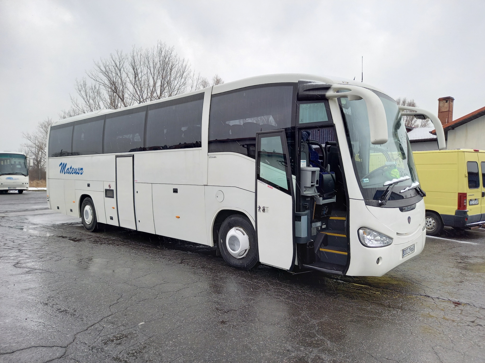 Biały autokar turystyczny marki Irizar z otwartymi drzwiami na mokrym parkingu, widoczny napis 'Mateusz' na boku pojazdu.