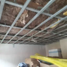 Sufit w trakcie ocieplania wełną mineralną, widoczna metalowa konstrukcja pod płyty gipsowo-kartonowe, fragment ściany i materiały budowlane w tle.