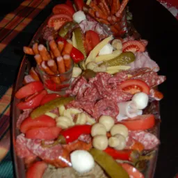 Dekoracyjna patera z wędlinami, serami, warzywami konserwowymi i świeżymi pomidorami, uzupełniona kabanosami w szklankach, na ciemnym tle.