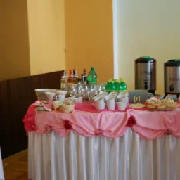 St&oacute;ł cateringowy z białym obrusem i r&oacute;żową dekoracją, na nim napoje w butelkach, szklanki, filiżanki, termosy z napisem 'Kawa' i 'Herbata' oraz przekąski.