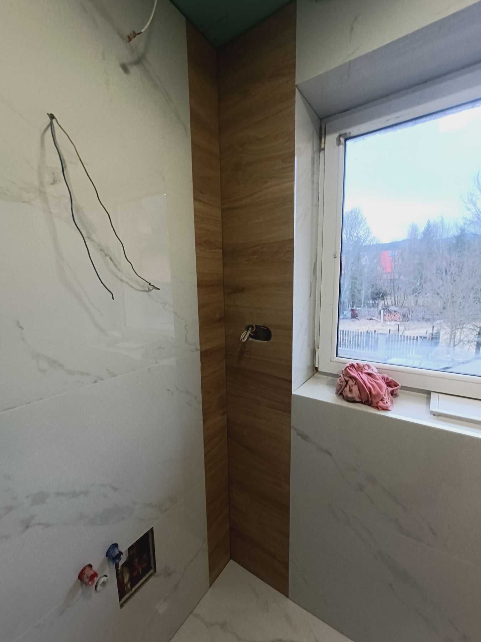 Wnętrze łazienki w trakcie wykańczania: białe płytki imitujące marmur, drewnopodobne panele ścienne, okno z widokiem na zewnątrz, wystające kable i instalacje.