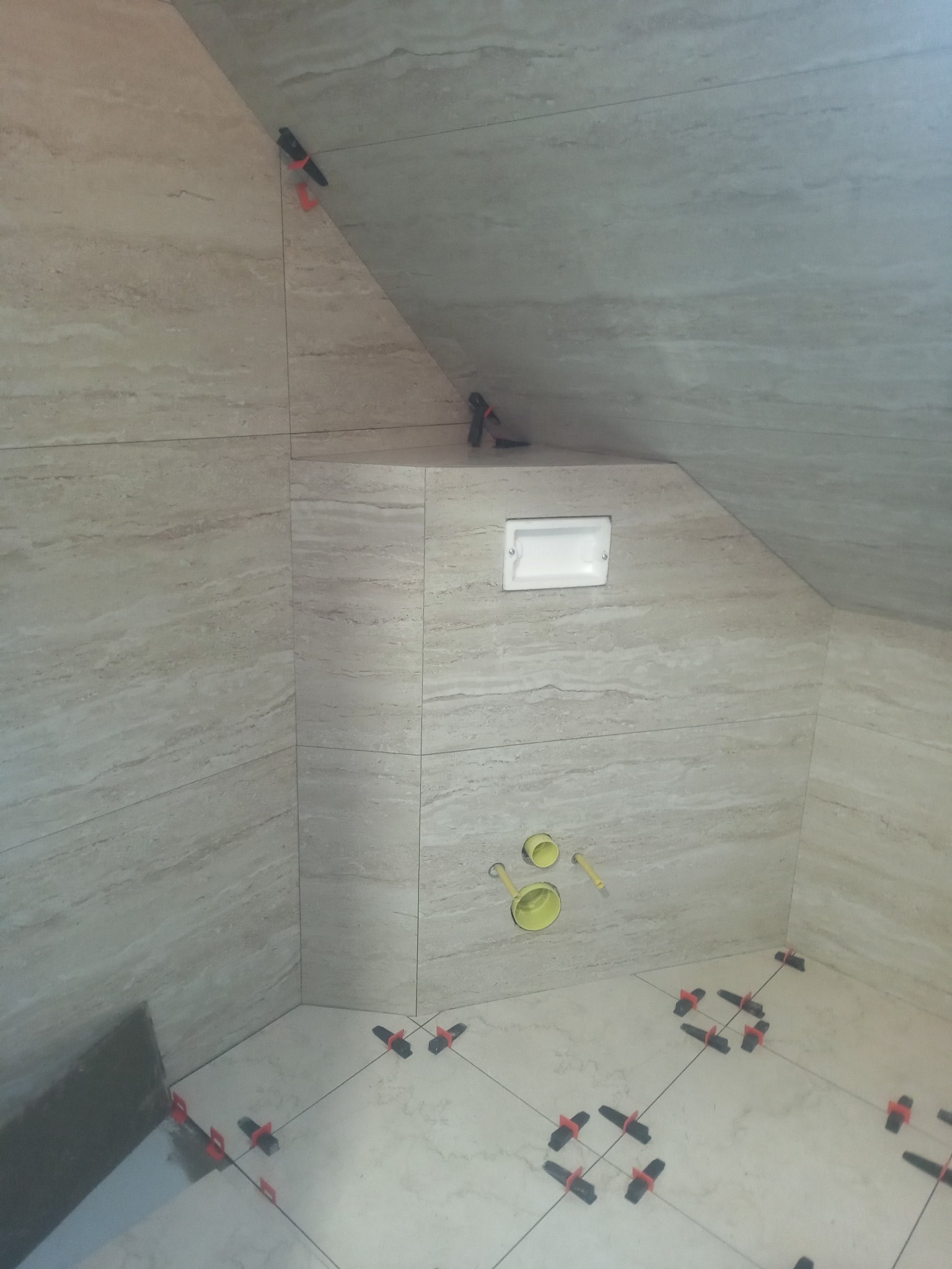 Układanie beżowych płytek imitujących marmur w łazience na poddaszu. Widoczne systemy poziomowania płytek i otwory na instalacje. Ukończona zabudowa geberitu.