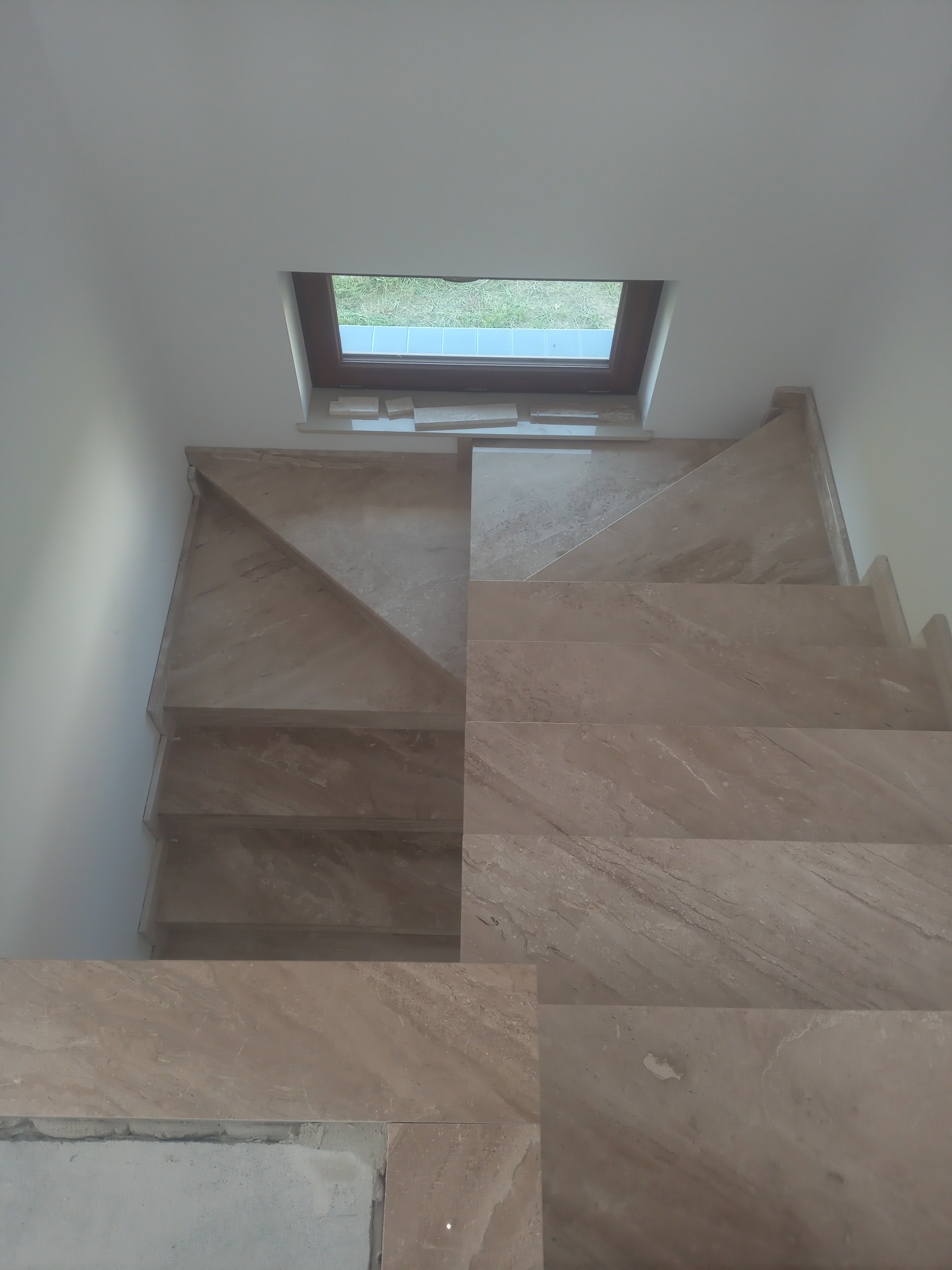 Wykończone schody wewnętrzne z beżowych płytek imitujących marmur, widok z góry, z oknem dachowym w tle. Częściowo widoczne niedociągnięcia przy dolnym stopniu.