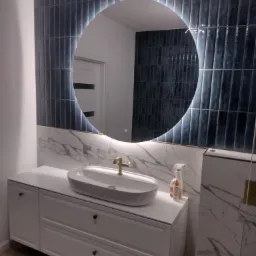 Nowoczesna łazienka z umywalką nablatową, baterią w kolorze złota, okrągłym podświetlanym lustrem i ciemnymi, pionowo ułożonymi płytkami na ścianie. Widoczne przewody elektryczne w górnym rogu...