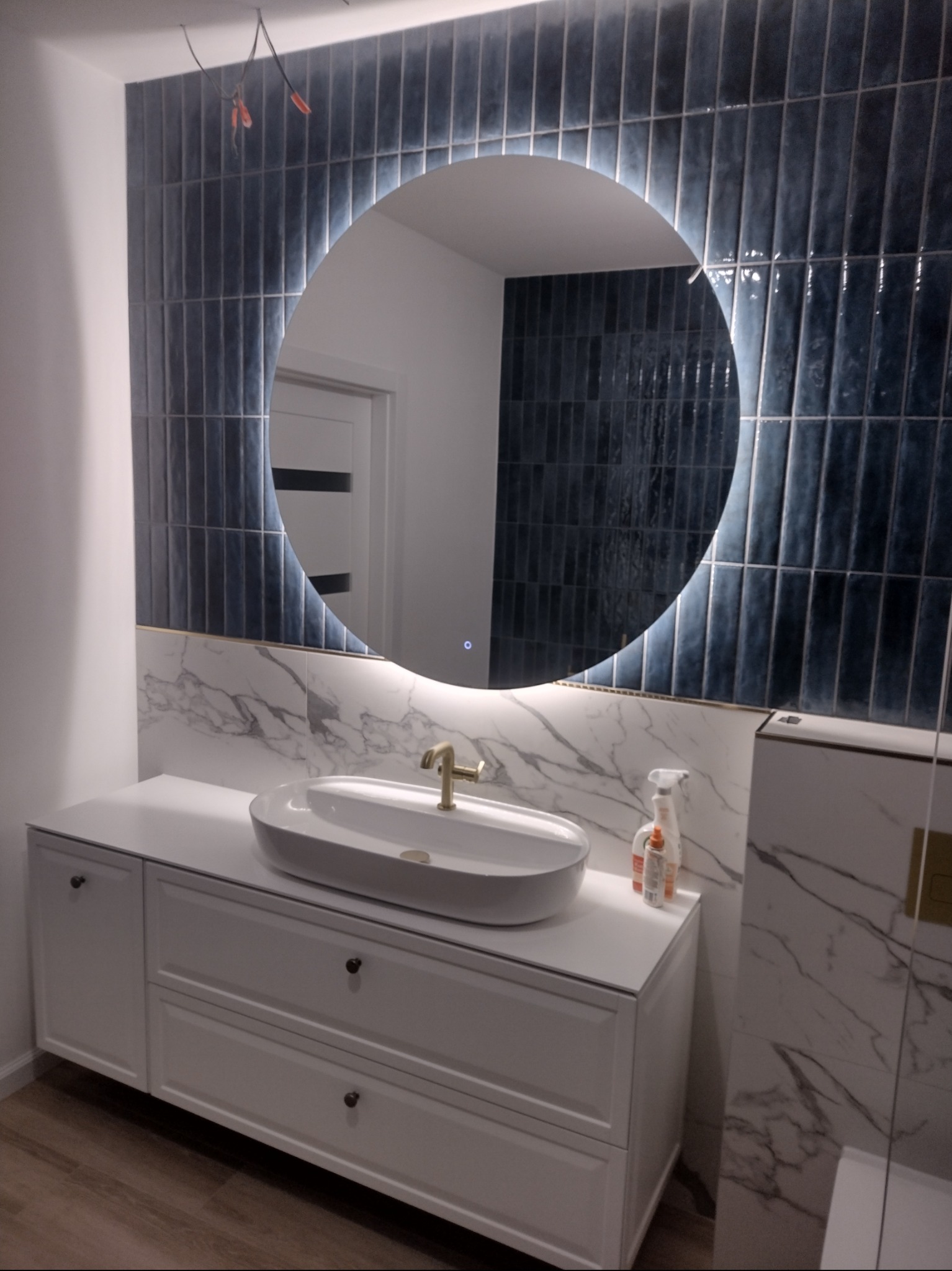 Nowoczesna łazienka z umywalką nablatową, baterią w kolorze złota, okrągłym podświetlanym lustrem i ciemnymi, pionowo ułożonymi płytkami na ścianie. Widoczne przewody elektryczne w górnym rogu...