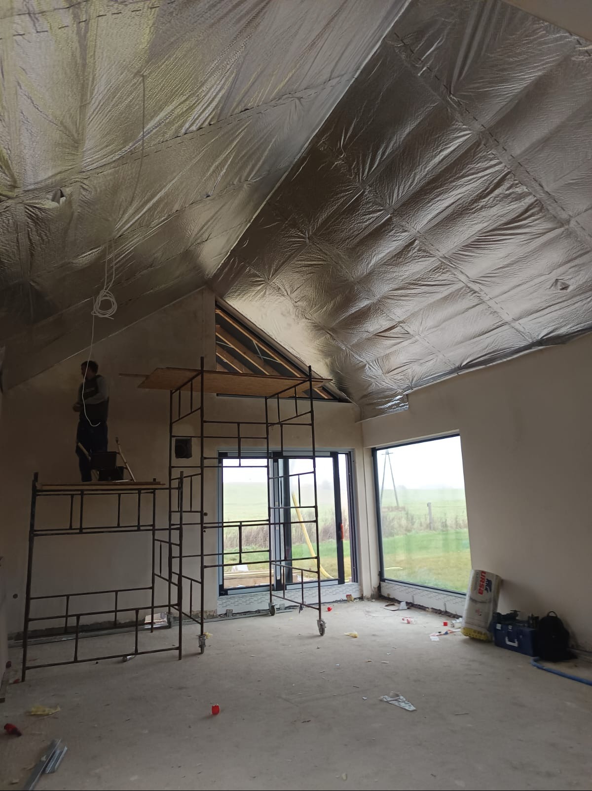 Prace wykończeniowe wnętrza: montaż izolacji termicznej na poddaszu, widoczne rusztowania, okna z widokiem na pole, worek z materiałem izolacyjnym, narzędzia pracy.