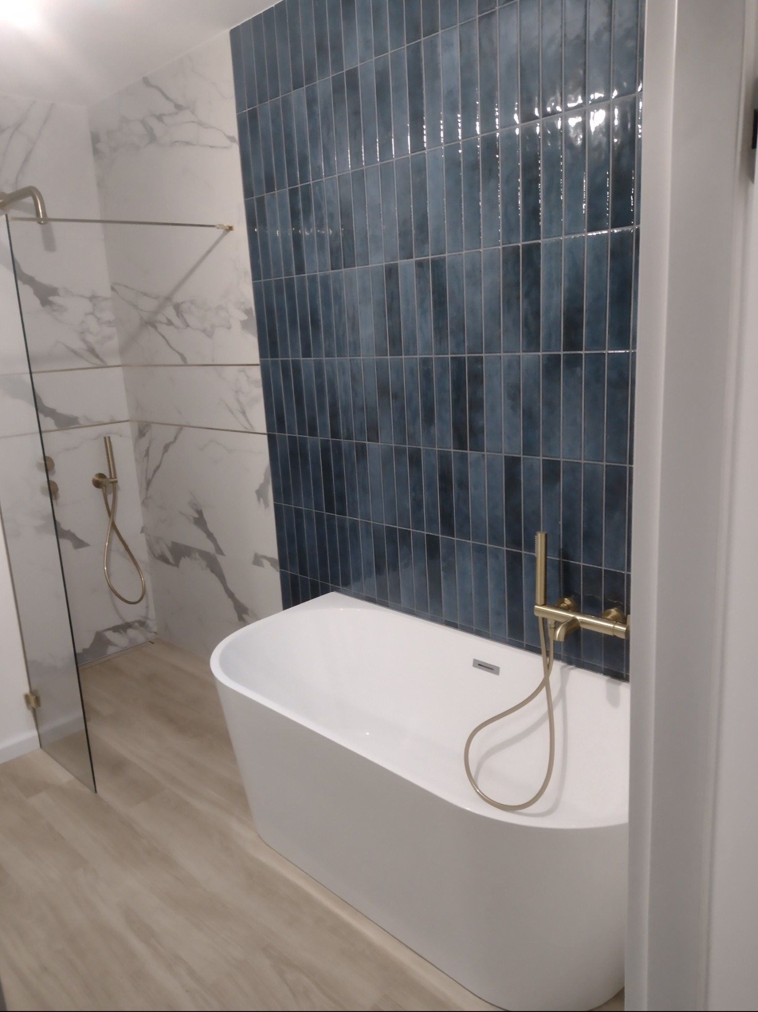 Nowoczesna łazienka z białą wanną wolnostojącą, baterią w kolorze złotym i ścianą wyłożoną pionowymi, niebieskimi płytkami. W tle widoczna kabina prysznicowa z marmurowymi płytkami.