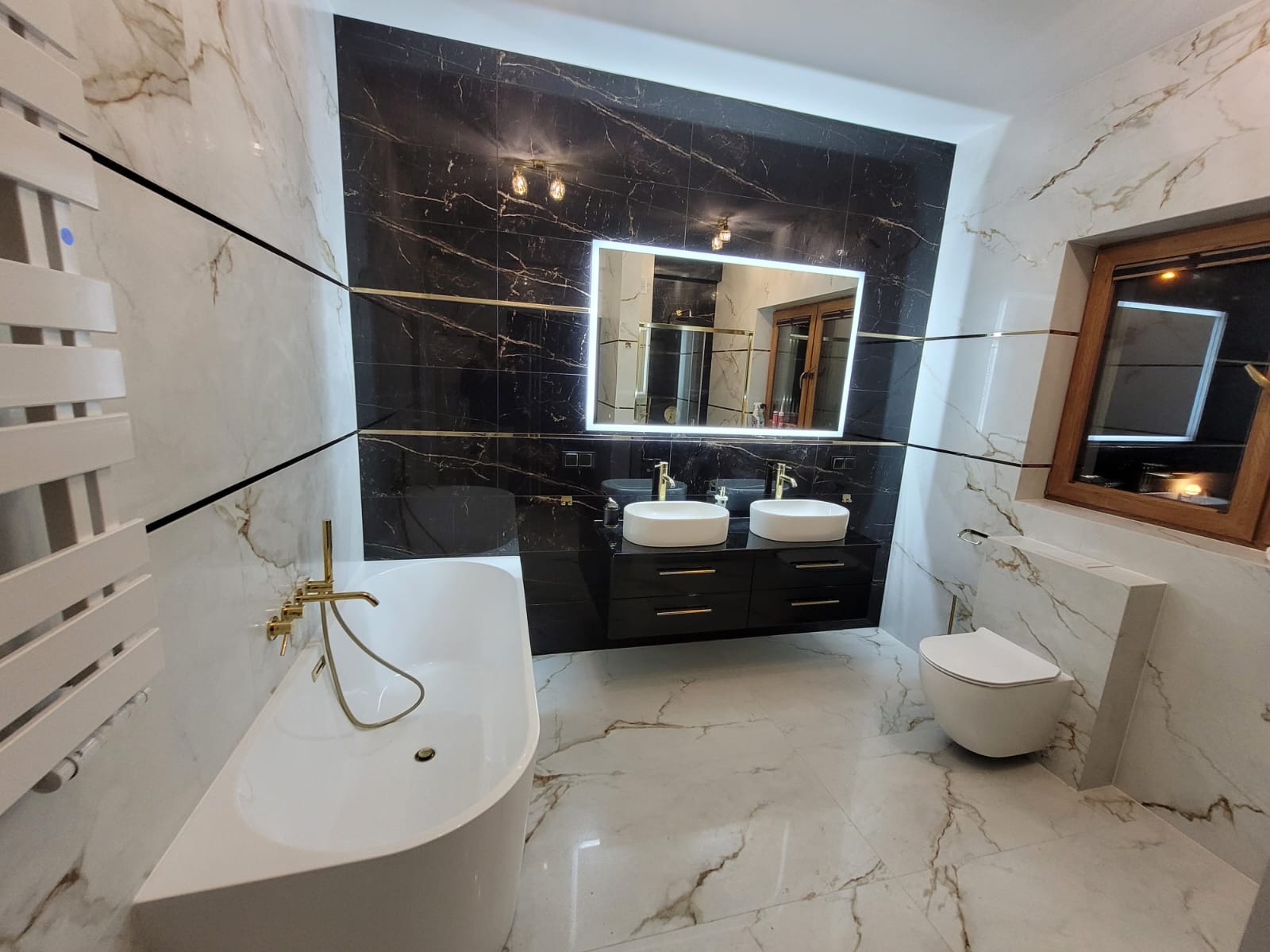 Nowoczesna łazienka z czarno-białymi marmurkowymi płytkami, podświetlanym lustrem nad czarną szafką z dwiema białymi umywalkami, wolnostojącą wanną z złotą armaturą i podwieszaną toaletą.