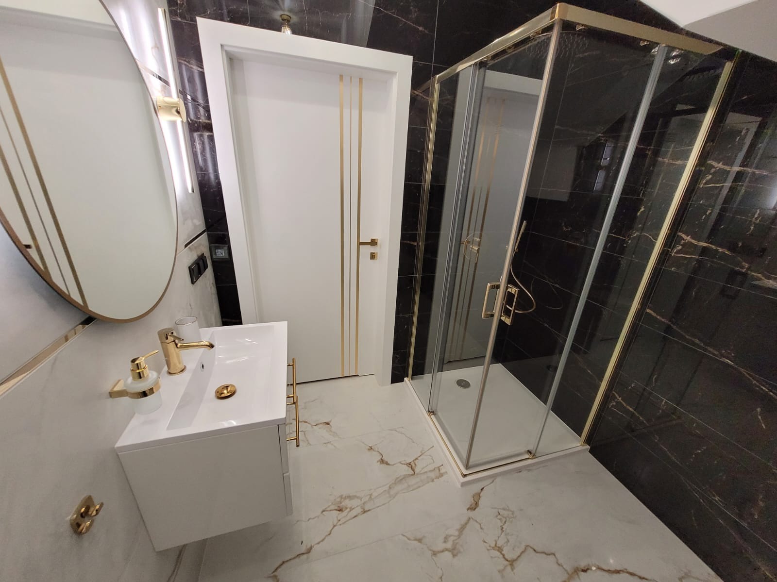 Nowoczesna łazienka z czarno-białymi marmurowymi płytkami, białą szafką umywalkową z złotą armaturą, kabiną prysznicową z złotymi ramami i białymi drzwiami z złotymi paskami.