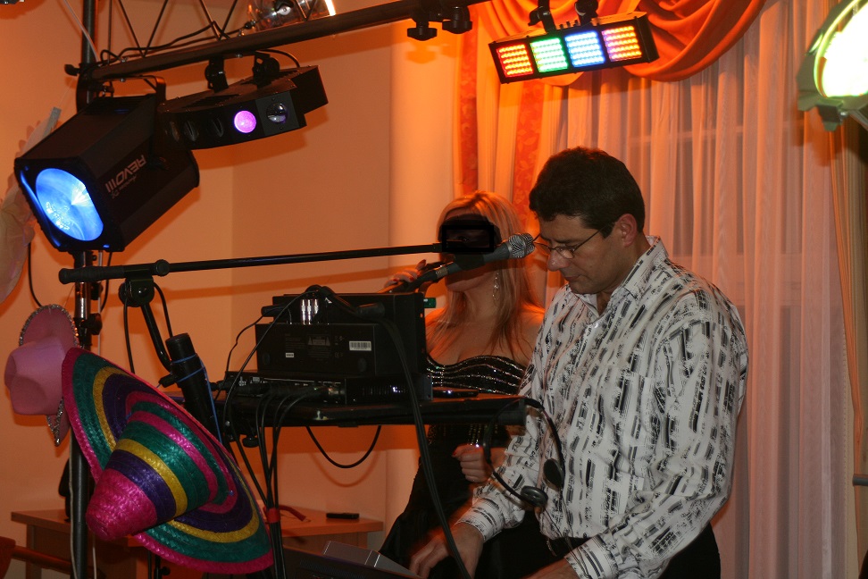 Muzyk grający na keyboardzie podczas występu, obok wokalistka przy mikrofonie, oświetlenie sceniczne i dekoracyjne kapelusze.