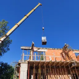 Żuraw budowlany podnosi pakiet materiałów na placu budowy domu w trakcie wznoszenia ścian, widoczne rusztowania i podpory, błękitne niebo w tle.